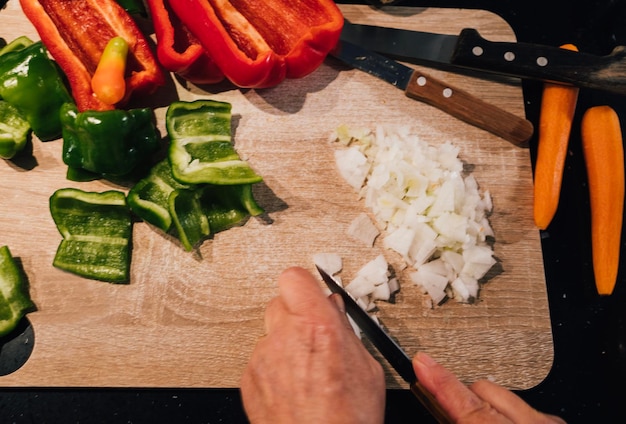 Vue de dessus des mains de femme coupant l'oignon sur une planche de bois avec des légumes rouges verts orange et blancs colorés autour