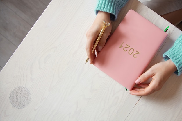Vue de dessus des mains de femme en pull chaud avec livre de journal 2021 couleur corail sur table. Projets futurs et réalisations pour la nouvelle année 2021. Bien-être