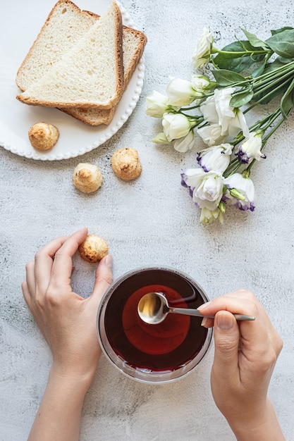 Vue de dessus des mains d'une femme avec une tasse de biscuits au thé, pain grillé et fleurs