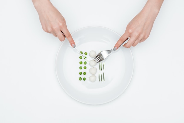 Vue de dessus des mains d'une jeune femme coupant et mangeant des pois verts, de la mozzarella et des herbes sur l'assiette