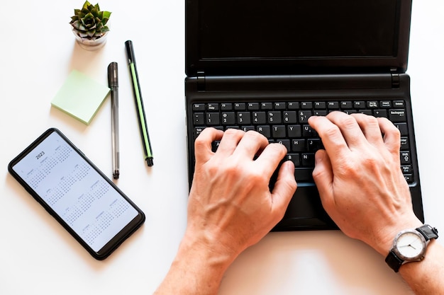 Vue de dessus des mains mâles tapant sur le clavier de l'ordinateur portable. Flatlay de bureau d'espace de travail blanc, téléphone portable