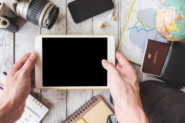 Photo vue de dessus des mains mâles tenant une tablette avec un équipement de voyage sur fond de table en bois blanc