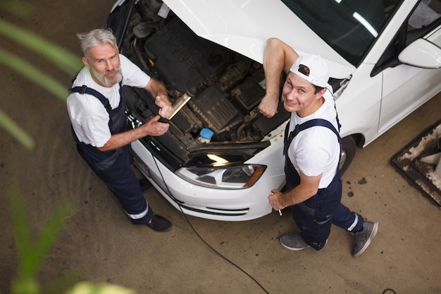 Vue de dessus d'un mécanicien automobile professionnel en salopette souriant à la caméra lors de la réparation d'une voiture