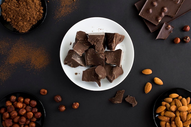 Vue de dessus des morceaux de chocolat noir sur plaque blanche poudre de cacao au chocolat et noix sur fond noir Fond de confiserie au chocolat