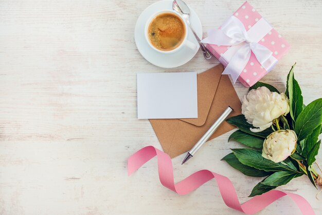 Vue de dessus de note vide, enveloppe kraft, tasse à café et fleurs de pivoine sur bois blanc rustique, fond.