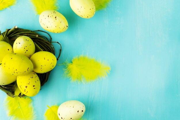 Vue de dessus d'un oeuf de Pâques jaune dans un nid de saule et des plumes jaunes sur fond turquoise texturé avec espace de message.