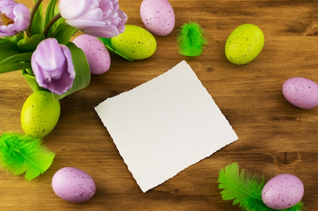 Vue de dessus d'un oeufs de Pâques colorés, tulipes violettes, plumes vertes et carte de message sur fond de bois brun.