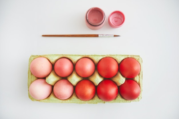 Vue de dessus des oeufs de Pâques rose et rouge pastel en caisse disposés en composition minimale avec pinceau sur fond blanc, espace copie