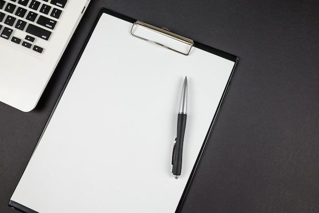 Vue de dessus d'un ordinateur portable blanc et d'un presse-papiers avec un stylo sur fond noir avec espace de copie