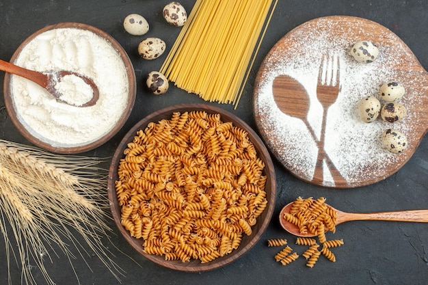 Vue de dessus des pâtes crues dans un bol brun et un couteau à fourche à pointes dessiné avec de la farine sur une planche à découper de la farine sur fond noir