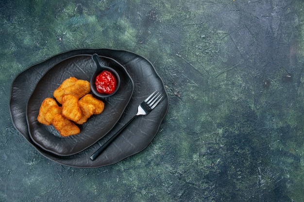 Vue de dessus des pépites de poulet et fourchette de ketchup dans des assiettes noires sur le côté droit sur une surface sombre