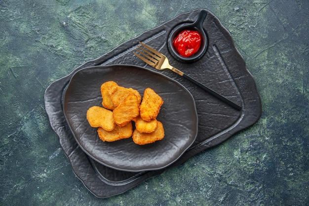 Vue de dessus des pépites de poulet sur une plaque noire et une fourchette de ketchup sur un plateau de couleur sombre