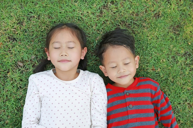 Vue de dessus Petit enfant asiatique garçon et fille allongé sur l'herbe verte à l'extérieur dans le parc d'été. Portrait de soeur et frère les yeux fermés se reposant dans le jardin heureux souriant ensemble