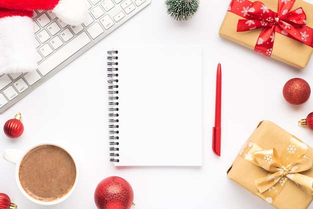 Vue de dessus photo de clavier tasse de boisson chaude chapeau de père Noël pin jouet boules de sapin de Noël rouge boîtes-cadeaux avec ruban arcs bloc-notes et stylo sur fond blanc isolé avec espace vide