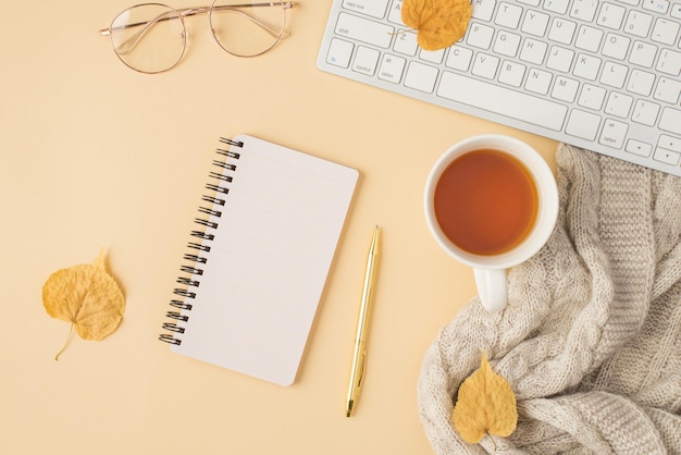 Vue de dessus photo du clavier de travail lunettes élégantes stylo doré cahier à spirale tasse de thé écharpe grise et feuilles d'automne jaunes sur fond beige isolé avec espace vide