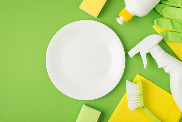 Vue de dessus photo d'un plat blanc propre au milieu vaporisateur de détergent et bouteilles de gel éponges brosse serviettes jaunes gants en caoutchouc vert sur fond vert isolé avec espace vide