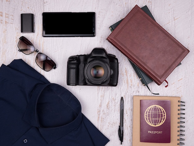 Vue de dessus à plat des accessoires de voyage sur fond de bois blanc. Appareil photo numérique, chemise, livres, smartphone, cahier papier avec stylo, lunettes de soleil et passeport