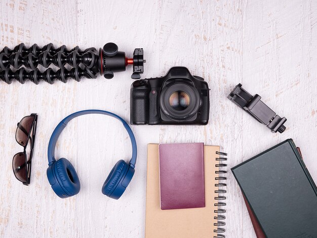 Vue de dessus à plat des accessoires de voyage sur fond de bois blanc. DSLR et caméra d'action, casque bleu, passeport, trépied, flash, ordinateur portable et smartphone