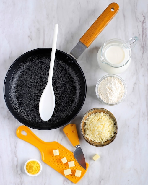 Vue de dessus poêle à frire, fromage, farine, lait, préparation d'ingrédients de sauce béchamel (sauce blanche pour pâtes) sur une table en marbre blanc