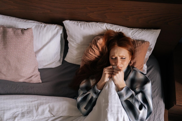 Vue de dessus portrait d'une jeune femme triste et déprimée pleurant se cachant sous une couverture allongée sur le lit dans une chambre sombre