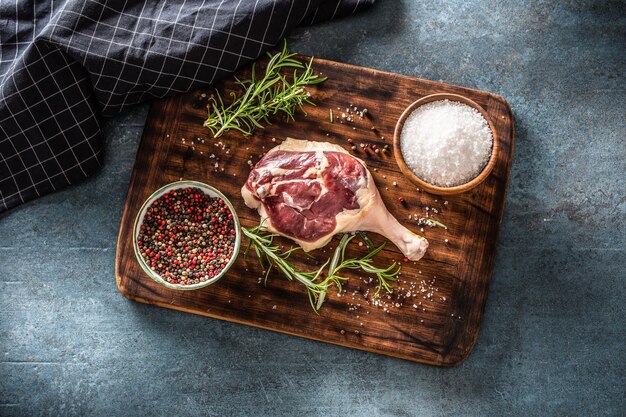 Vue de dessus de la préparation de viande dans un environnement rustique sombre. Cuisse de volaille non cuite, romarin frais, poivre et sel placés sur une planche à découper marron foncé à côté d'un chiffon.