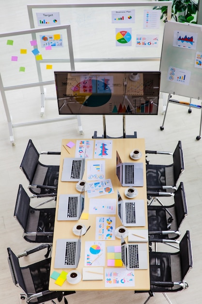 Vue de dessus de la salle de réunion de conférence dans le bureau de l'entreprise pleine d'ordinateurs portables sur un bureau avec des chaises noires et un moniteur grand écran et un tableau en verre avec des documents papier de données.