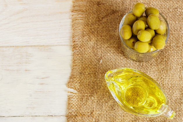 Photo vue de dessus de la saucière en verre avec de l'huile d'olive extra vierge et des olives vertes fraîches sur une toile de jute sur une table en bois.