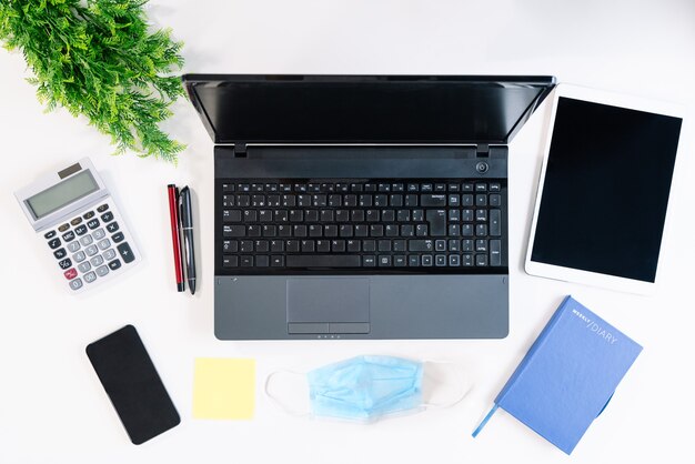 Vue De Dessus D'une Table De Bureau Blanche Avec Un Ordinateur Portable, Une Calculatrice, Un Téléphone Portable, Un Poster, Des Stylos, Un Ordinateur Portable, Une Tablette Et Un Masque Facial En Raison De La Pandémie De Coronavirus Covid19, Le Tout Sous Un Très Bon éclairage