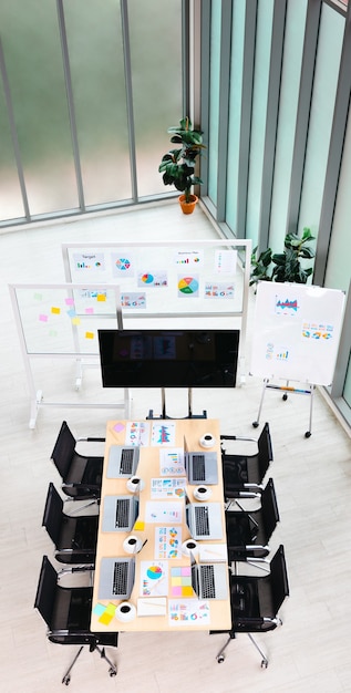 Vue de dessus de la table de travail de la salle de réunion dans le bureau vide de l'entreprise, plein d'ordinateurs portables, des tasses à café, des documents de documents de données, des chaises noires et un panneau de verre à écran vierge près des fenêtres.