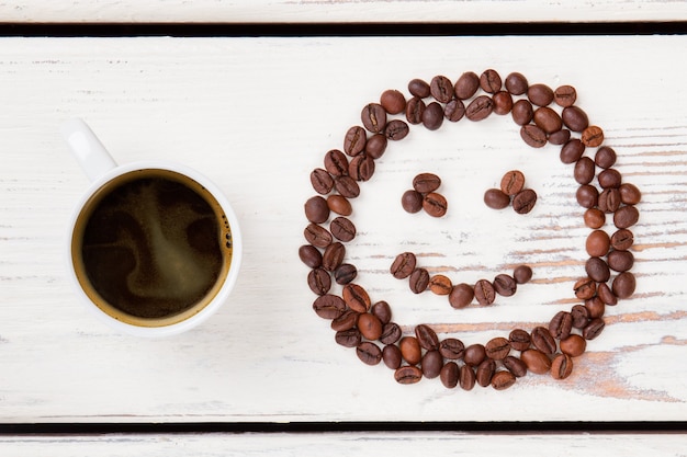 Vue de dessus tasse de café chaud et haricots en forme de visage souriant. Planches de bois blanches en surface.