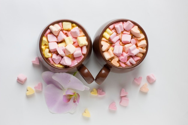 Vue de dessus des tasses brunes de chocolat chaud avec des guimauves en forme de coeur sur fond blanc Fond romantique avec chocolat chaud et fleur d'orchidée Concept d'amour pour la Saint-Valentin ou le mariage