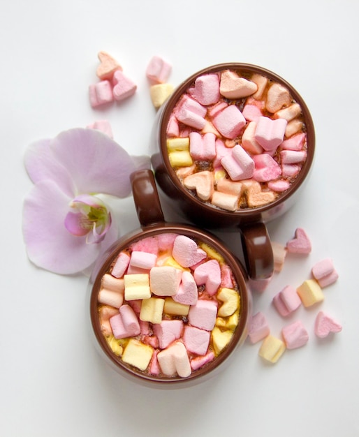 Vue de dessus des tasses brunes de chocolat chaud avec des guimauves en forme de coeur sur fond blanc Fond romantique avec chocolat chaud et fleur d'orchidée Concept d'amour pour la Saint-Valentin ou le mariage