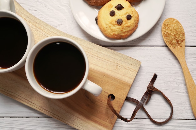 Vue de dessus des tasses de café chaud avec une cuillère de sucre et de biscuits aux pépites de chocolat sur une plaque en bois