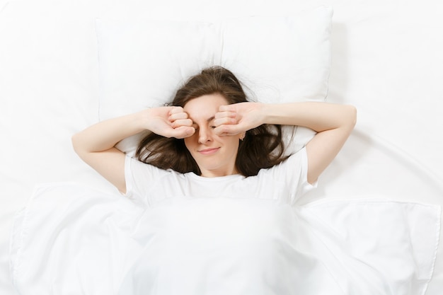 Vue de dessus de la tête d'une jeune femme brune heureuse allongée dans son lit avec un drap blanc, un oreiller, une couverture. Femme souriante s'étirant dans son lit, passant du temps dans la chambre