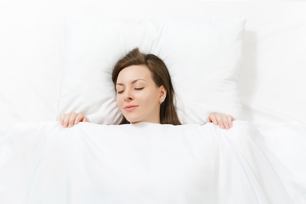Vue de dessus de la tête d'une jeune femme brune heureuse allongée dans son lit avec un drap blanc, un oreiller, une couverture