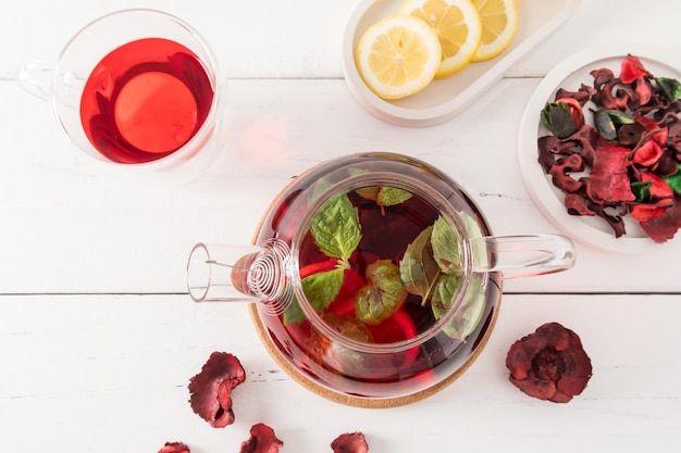 Vue de dessus de la théière avec du thé aux pétales de rose d'hibiscus fraîchement infusé et des feuilles de menthe au citron pour améliorer le goût et les vitamines table en bois blanc