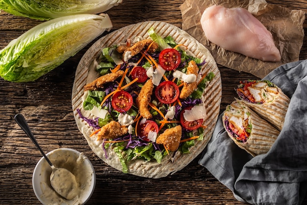 Vue de dessus d'une tortilla ouverte avec des pépites de poulet, des tomates cerises, du chou rouge, de la salade, des rayures de carottes et des copeaux de parmesan.