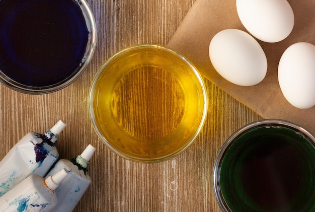 Vue de dessus de trois bols avec de l'eau colorée pour colorer les œufs en bleu, jaune, vert et œufs blancs sur fond en bois et tubes de colorant alimentaire