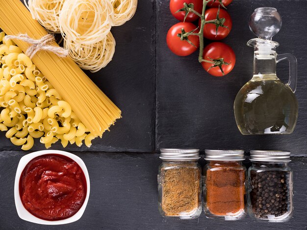 Vue de dessus variété de pâtes crues non cuites à côté d'un bol avec de la sauce tomate, des tomates fraîches, différentes épices et une bouteille d'huile de tournesol