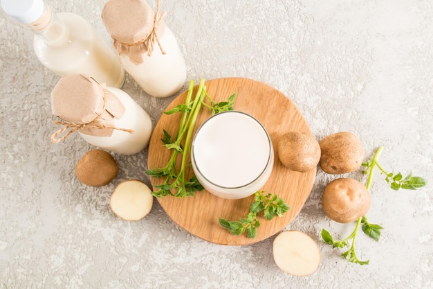 Vue de dessus d'un verre rempli de lait de pomme de terre biologique sur une planche de bois ronde deux bouteilles de tubercules de lait et de feuilles sur une table en béton