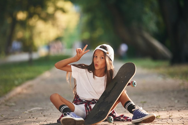 Photo vue de devant montrant des gestes une petite fille heureuse avec un skateboard à l'extérieur