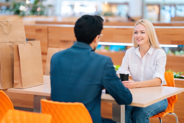 Vue de dos à l'homme méconnaissable et à la jolie femme blonde ayant un rendez-vous assis à table se regardant dans le centre commercial. Beau jeune couple buvant du café et parlant au café après le shopping.