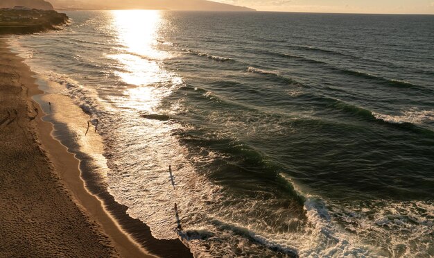 Vue de drone de personnes sur une plage de sable lavée par des vagues mousseuses de mer sous un soleil radieux au coucher du soleil