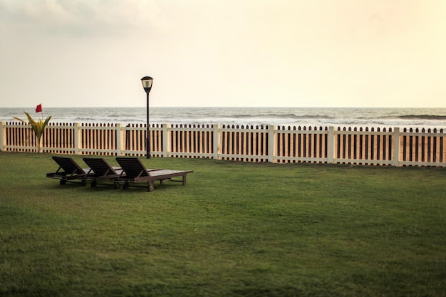 Vue du complexe hôtelier sur les transats vides sur la pelouse verte, la plage et la mer derrière une clôture en bois après le coucher du soleil.