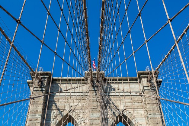 Vue du pont de Brooklyn avec haubans diagonaux et câbles de bretelles verticales. Au centre de la photo se trouve le drapeau américain flottant au vent.