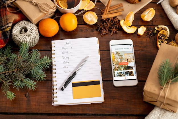 Vue du smartphone avec promo, bloc-notes avec stylo, liste et carte de crédit entouré de trucs de Noël traditionnels sur la table