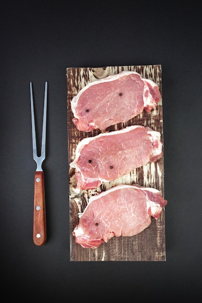 Vue élevée des steaks de viande crue alignés avec des épices et une fourchette à viande sur une planche à découper de cuisine sur fond sombre