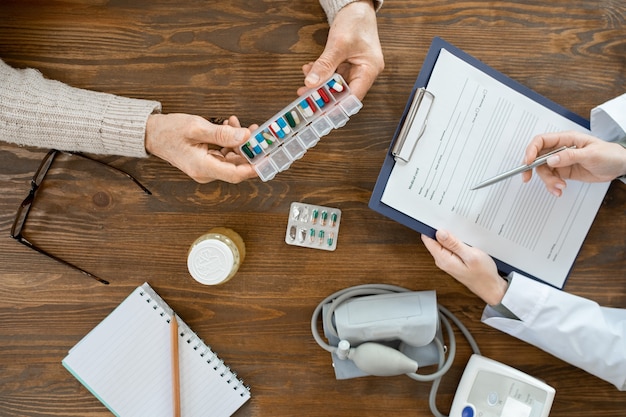 Vue d'ensemble des mains d'un homme âgé malade tenant un récipient avec des pilules sur une table lors d'une consultation médicale avec un médecin remplissant le document