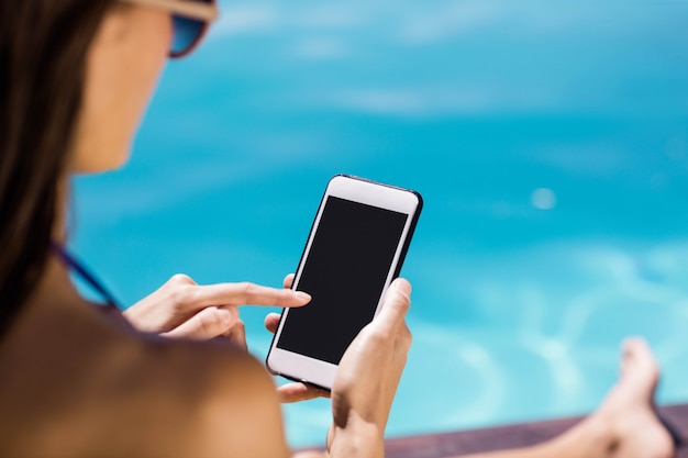 Vue sur l&#39;épaule de brune à l&#39;aide de smartphone au bord de la piscine