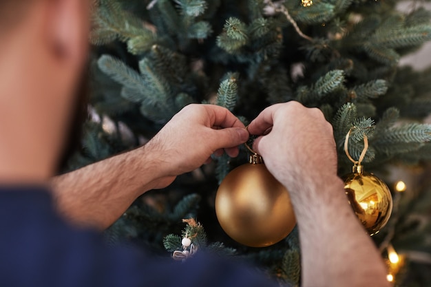 Vue d'épaule d'un homme suspendu à une boule de noël dorée sur un arbre tout en se préparant à la célébration